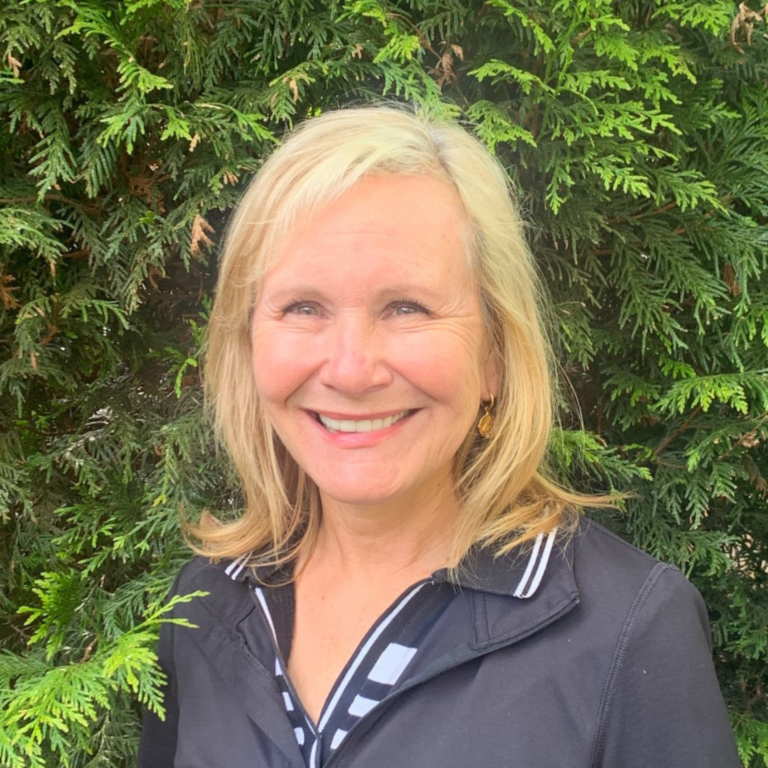 Bea Hadikin smiles outside while standing in front of green hedges. She is wearing a black coat and has shoulder length blond hair.