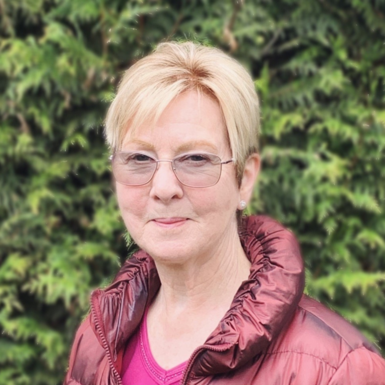 Marie Sabine smiles outside in front of a green hedge. She wears glasses, has show blond hair, and wears a pink shirt with a red jacket over top.