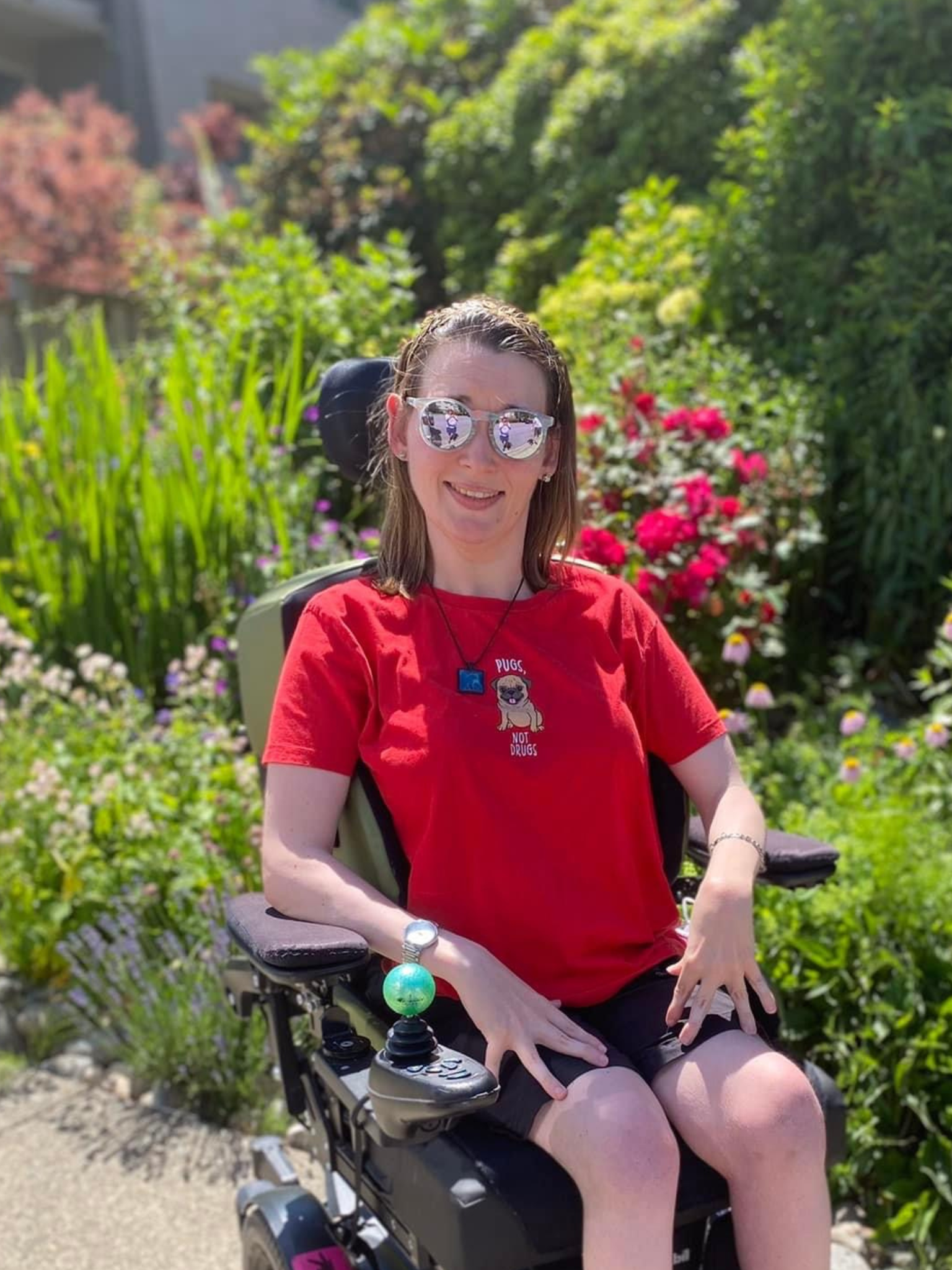 Madison Van Oene poses for a photo outside in a garden. She uses a powered wheelchair and wears a red shirt, black shorts and sunglasses. She has shoulder length, brown hair.