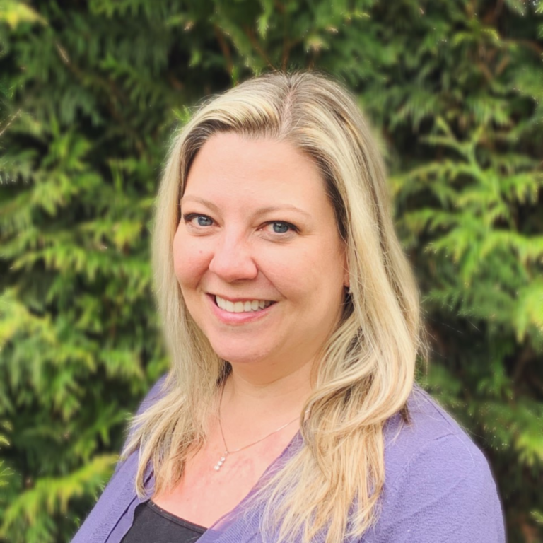 Nicole Russell smiles outside in front of green hedges. She wears a purple cardigan over a grey top. She has long blond hair.