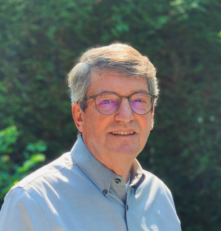 Ian Jarvis stands in front of a green hedge. He has short grey hair, glasses and wears a buttoned up, collared dress shirt.