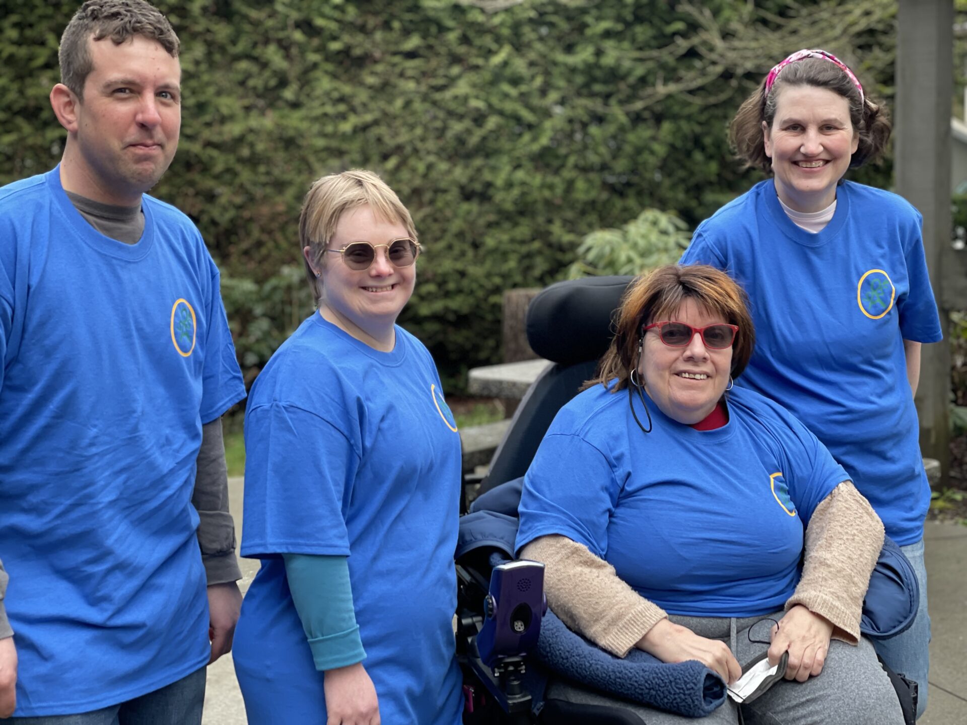 4 members of SAS wearing blue shirts, standing outside. 3 of them are standing and 1 of them is in a motorized wheelchair.